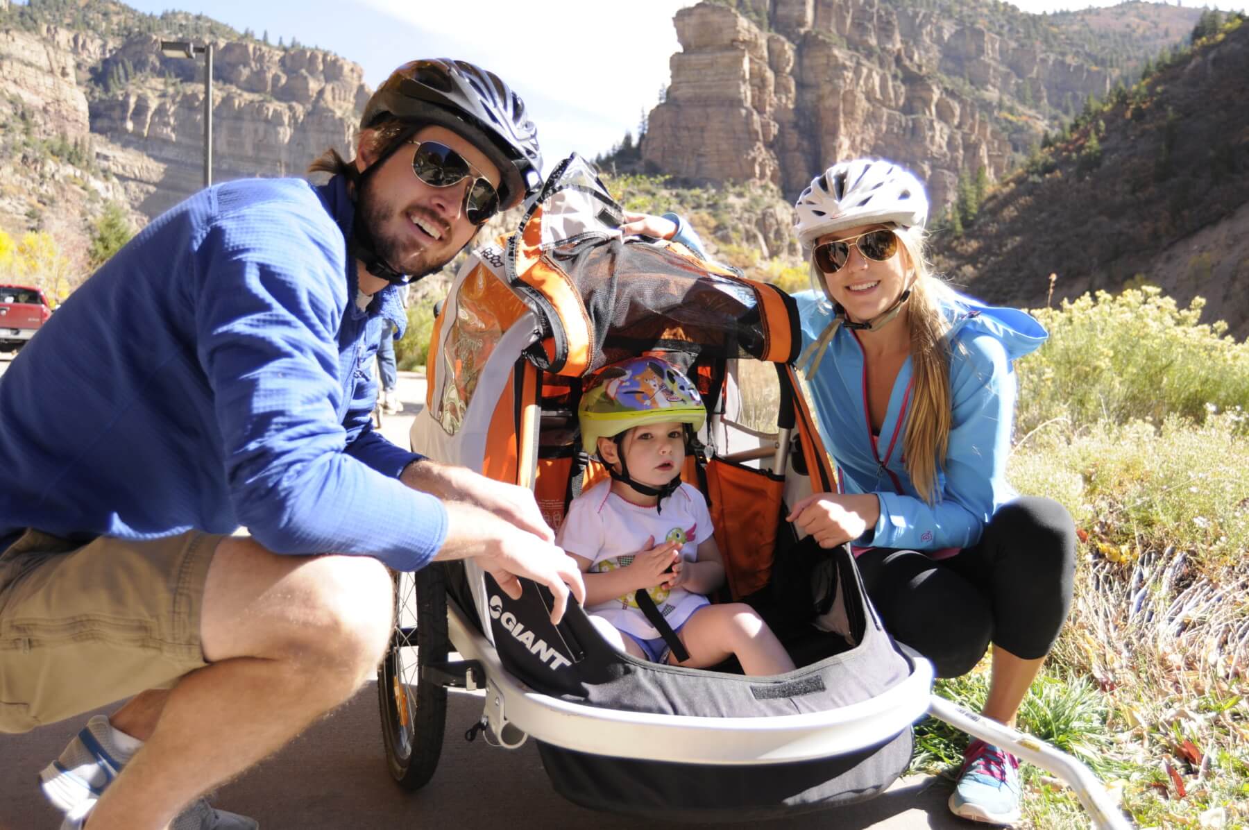 A photo of three people on a bike trail in Glenwood Canyon. There are two adults posing next to a kid in a bike trailer.