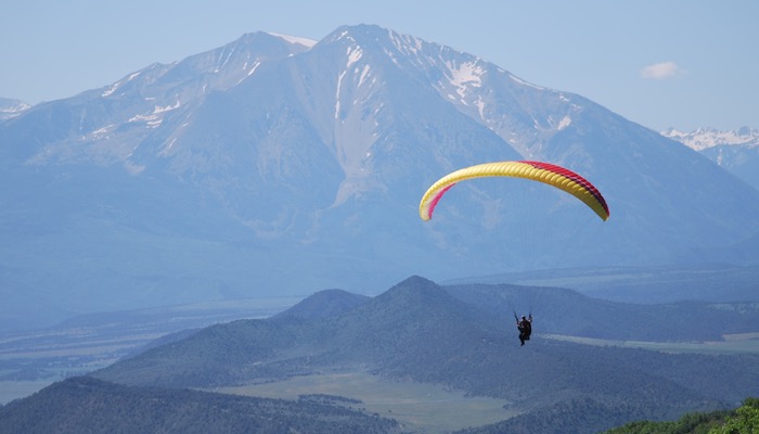 A photo capturing a lone paraglider soaring above Glenwood Springs with the scenic backdrop of Mt. Sopris on a hazy, yet blue-sky day.