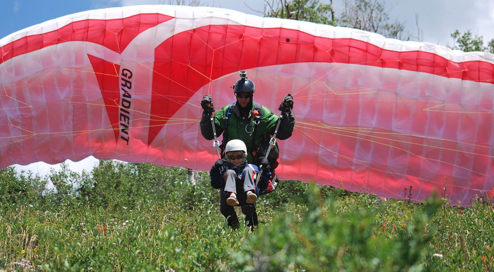 Capturing the essence of a tandem paragliding adventure, this photo showcases two paragliders gracefully descending to land in a lush green field. Both individuals are equipped with helmets, sunglasses, pants, and closed-toed shoes, emphasizing safety and preparedness for the landing. The vibrant green surroundings and the synchronized descent of the tandem paragliders create a harmonious scene of exhilaration and shared airborne excitement.