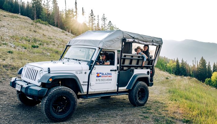 This delightful image captures a white Blazing Adventures jeep, transporting a joyous family on a mountain jeep tour. Radiant smiles adorn every face as the four-wheel drive vehicle navigates the scenic terrain. The shared enjoyment and excitement of the ride are palpable, creating a memorable and heartwarming moment during their mountain adventure with Blazing Adventures. The sun shines through the trees.