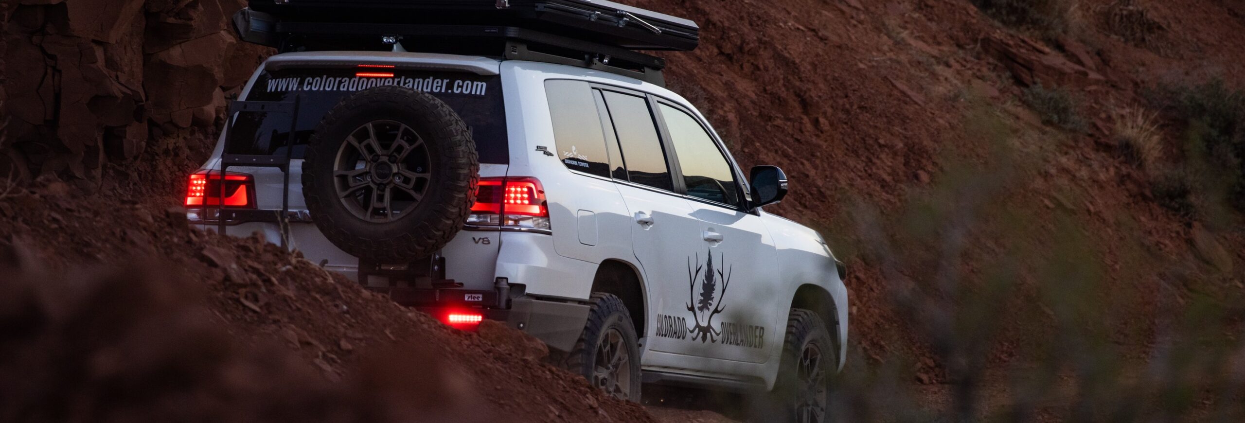 In this close-up photo, Colorado Overlander's Land Cruiser confidently navigates a vibrant red dirt road. The details are crystal clear, revealing the distinct logo on the vehicle and the rooftop tent, emphasizing the rugged and adventurous spirit embodied by this exploration-ready setup.