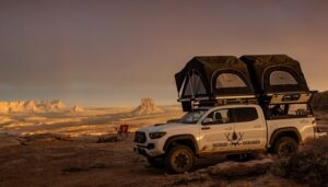 In this striking image, a Colorado Overlander truck is showcased atop a mountain with its rooftop tents open, providing a breathtaking vantage point overlooking a dramatic desert scene.