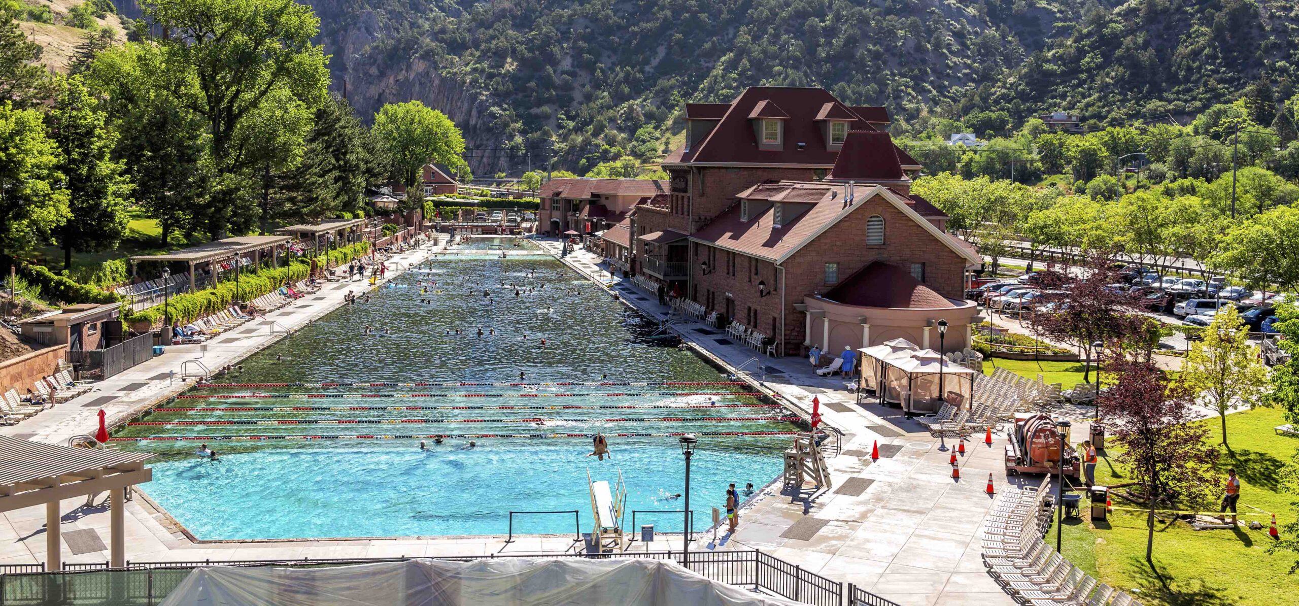 This photo captures the serene charm of Glenwood Hot Springs pool as seen from the pedestrian bridge in Glenwood Springs during the day. In the backdrop, the green mountains with scattered dirt patches add a rustic touch to the natural beauty of the scene.