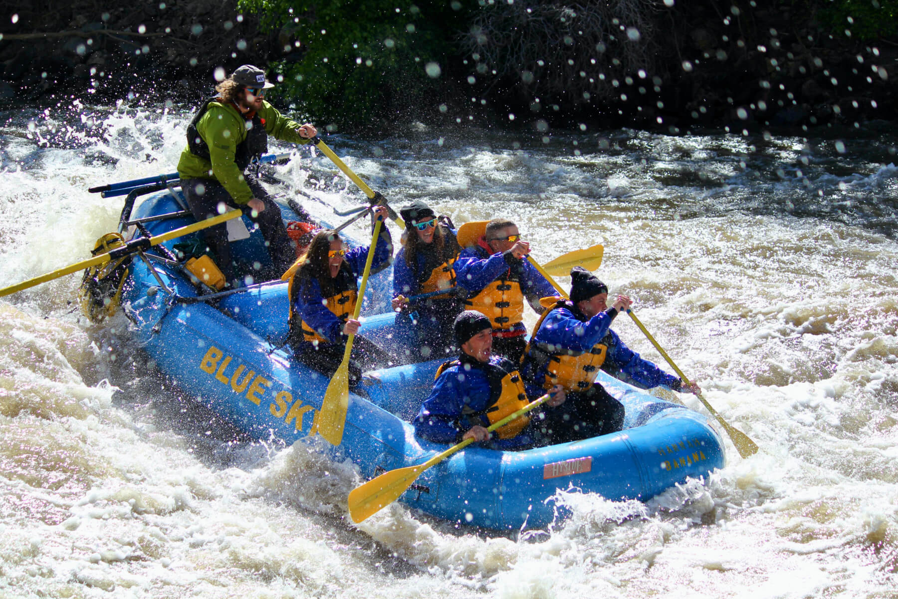 An image capturing the adventure of a Blue Sky-branded raft navigating the Shoshone Rapids on the Colorado River. The excitement is palpable, marked by joyous smiles. The dynamic scene is intensified by water splashes that surround the raft, creating a visually striking and exhilarating composition.