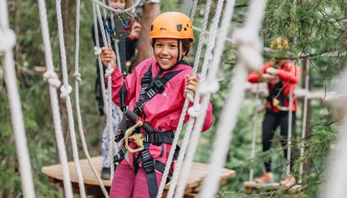 This image captures a girl in a harness conquering the exhilarating adventure course of the Lost Forest in Snowmass Village, CO. Suspended in mid-air, she holds the rope and balances for the photo, navigating the challenges with courage and determination. She is surrounded by the scenic beauty of the forest.