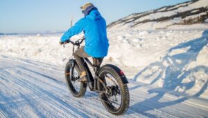 Embark on a winter ride with this striking image featuring a person riding a black fat tire e-bike through the snowy terrain. Clad in a blue ski jacket, dark pants, and boots, the rider is geared up for the cold adventure. The figure is also wearing a hat and sunglasses, navigates the winter wonderland with style and determination.