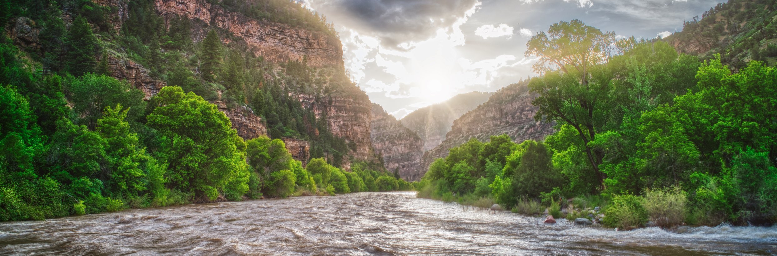 This serene photograph beautifully captures the calm Class II waters of the Colorado River. The surroundings unfold with a perfect view of canyon walls and lush vegetation embracing the river, creating a tranquil and picturesque scene. The radiant sun takes center stage, adding warmth and radiance to the peaceful waterscape.
