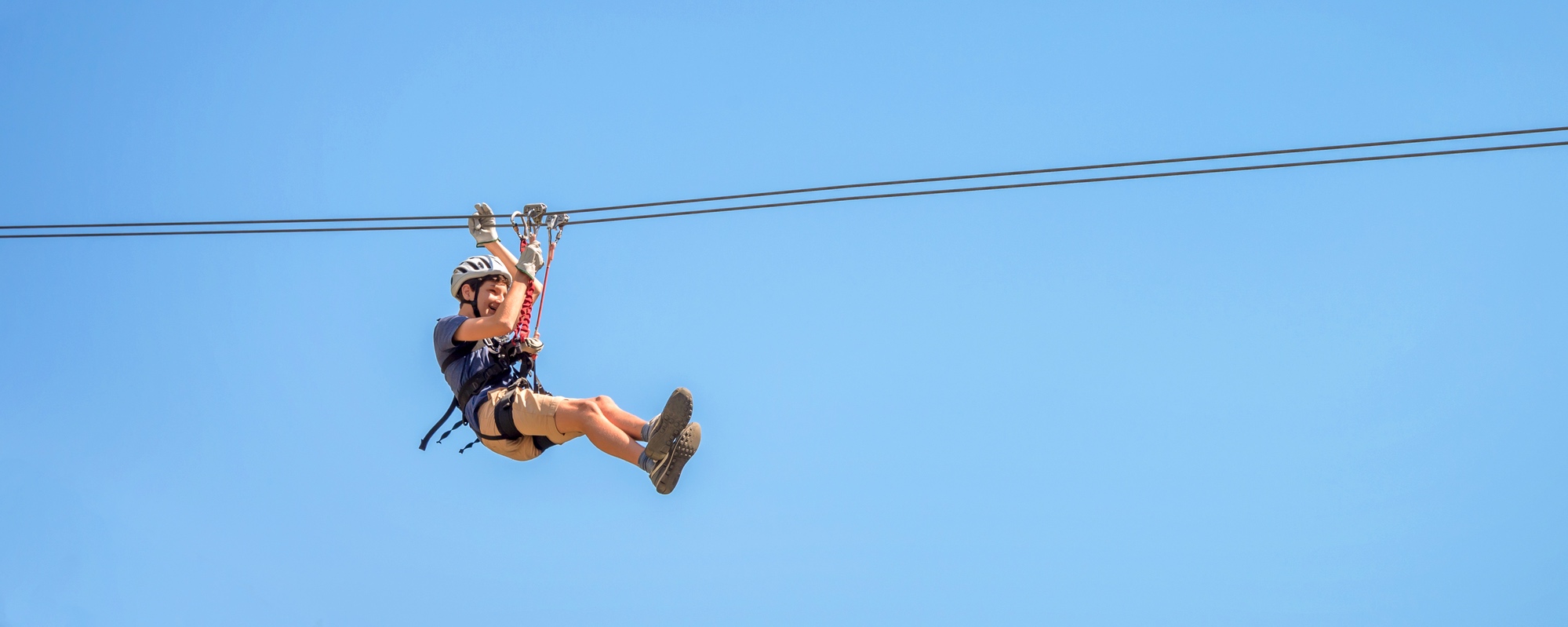 This is of an image of a single zipliner, fully geared up, soaring through the air against a cloudless blue sky backdrop.