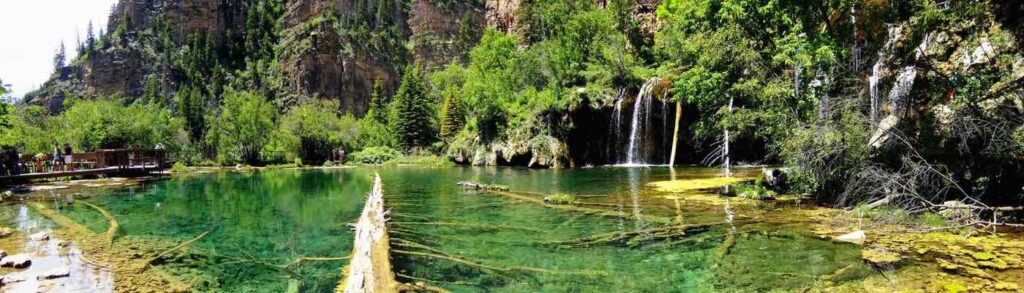 This is a photo of Hanging Lake in Glenwood Canyon. The colors around the lake are extremely vibrant with bright shades of green. A wooden observation deck overlooks the clear, teal waters for visitors' viewing pleasure.