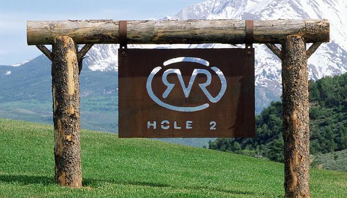 In this image, a sign marking Hole 2 at River Valley Ranch is charmingly nestled between two tree trunks, creating a rustic aesthetic against the mountainous backdrop. The scene blends the rustic charm of nature with the structured elements of the golf course.