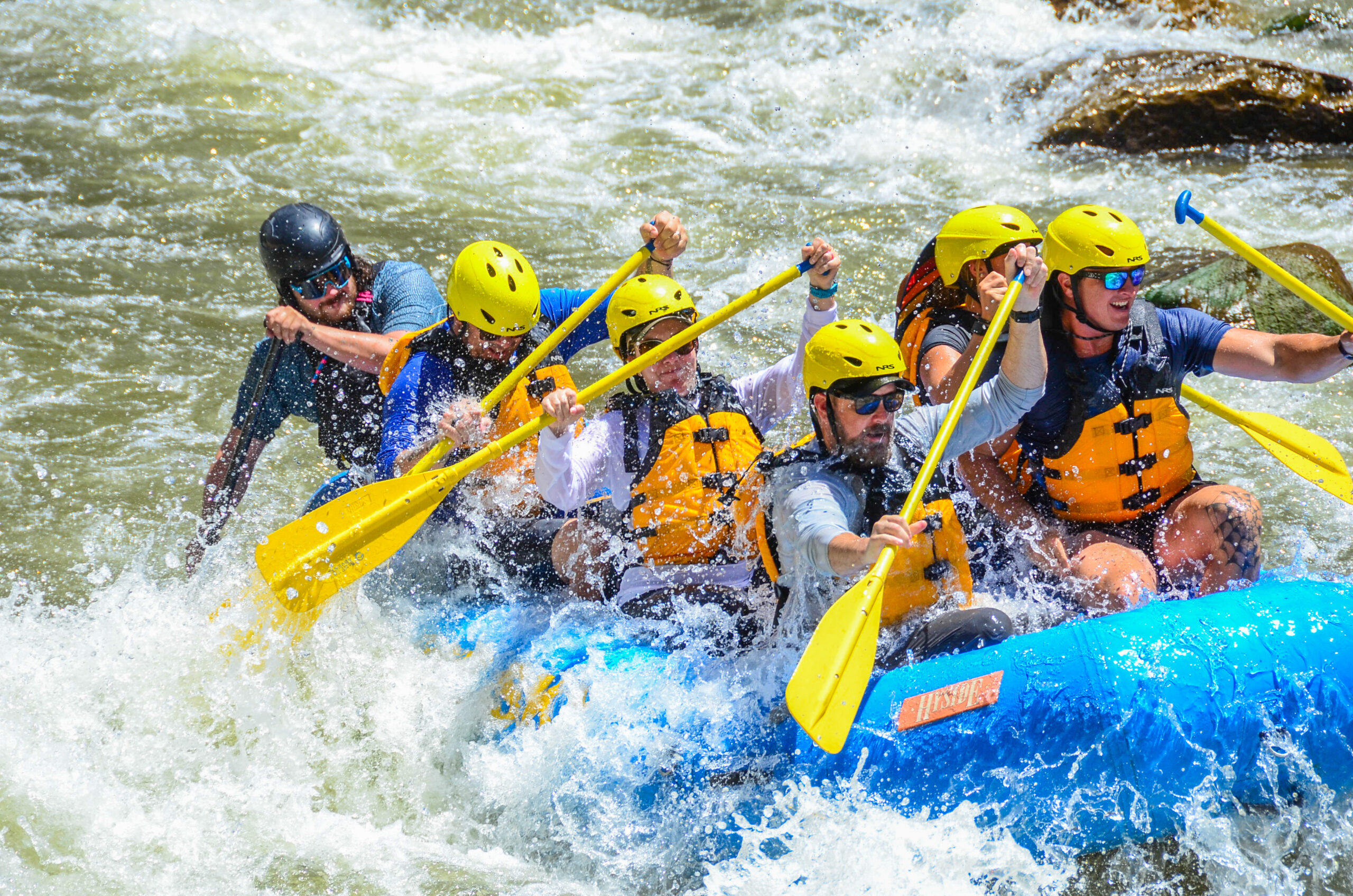 In this compelling image, a proficient guide expertly directs a fully occupied boat from the rear as participants paddle through the whitewater together. Cloaked in life jackets and helmets, the group showcases seamless teamwork, with water splashing around them, capturing the dynamic and exhilarating essence of their adventure.