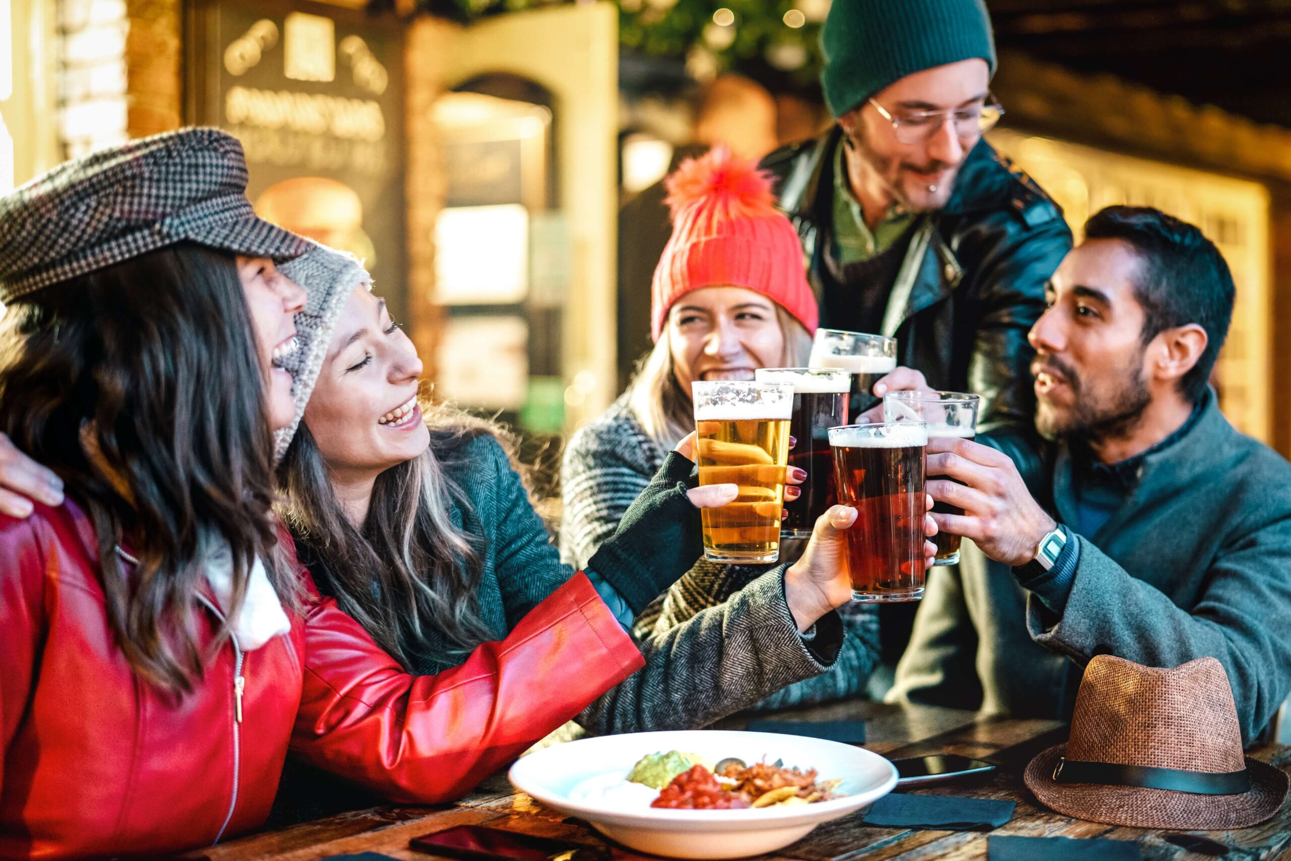 A lively group of friends raises their beers in a cheerful toast over a plate of food. Their vibrant winter attire adds to the festive atmosphere, and the big smiles on their faces capture the joy and excitement of their social gathering.