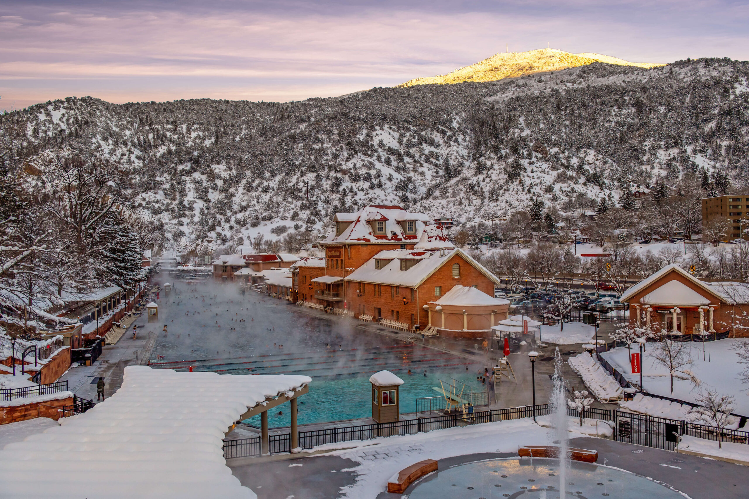 Capture the winter beauty of Glenwood Hot Springs pool from the pedestrian bridge, framed by snow-covered mountains. The tranquil scene unfolds as soothing hot springs mineral waters release steam, creating a serene contrast against the snowy landscape.