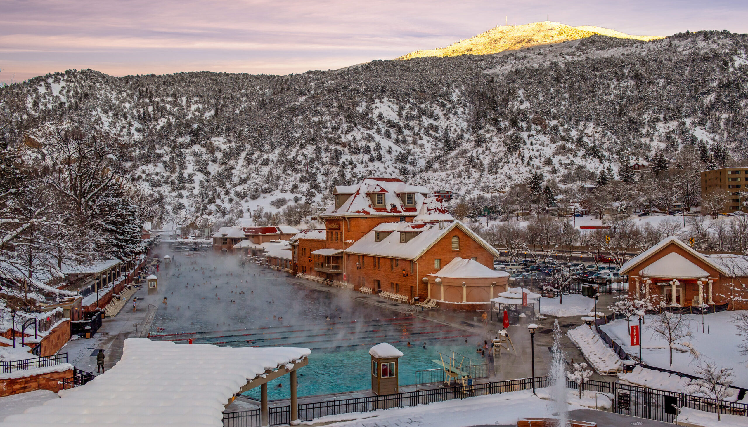 This image shows the hot springs pool during the winter time at sunset. You can see the sun setting on the mountain in the background. The steam from the hot springs pool can be seen with snow in all the surrounding buildings and areas.