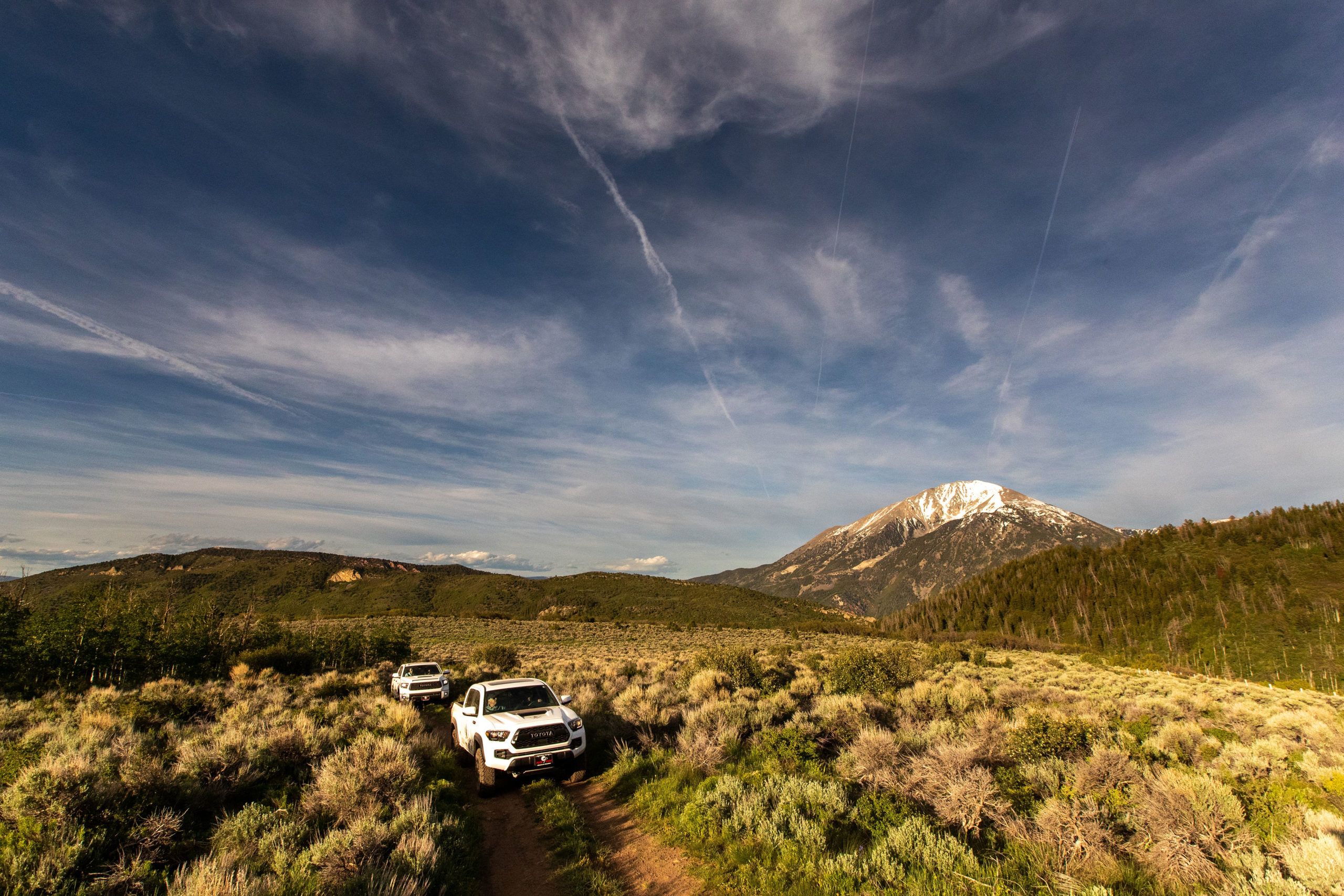 Two vehicles traverse a scenic dirt road winding through the Colorado backcountry, surrounded by sage bush. The picturesque scene exudes solitude, creating an impression that they are the sole adventurers in the vast and beautiful landscape.