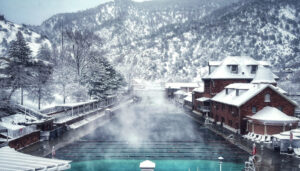 Scenic winter view of Glenwood Hot Springs pool, surrounded by snow-covered mountains and buildings. The soothing hot springs mineral waters create a tranquil scene with steam rising, offering a serene contrast to the snowy landscape.