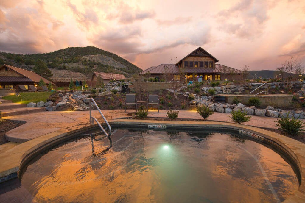 In this photograph, the hot pool at Iron Mountain Hot Springs Resort takes center stage, creating a serene and tranquil atmosphere. The view extends back to the main office and bathhouse, with subtle orange and white clouds gracing the sky above. The image captures the peaceful essence of this inviting retreat.