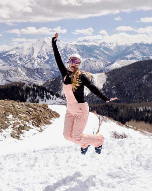 In this exuberant image, a woman leaps for joy against the backdrop of the mountains. Clad in her winter ski gear, complete with goggles and a helmet, she radiates pure excitement, smiling broadly with her hands raised in the air.