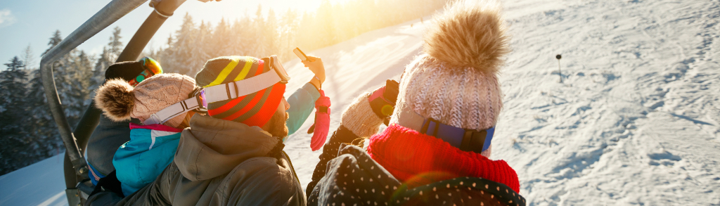 A group of friends enjoys a scenic ride on the chairlift, overlooking the ski run below and the picturesque mountain trees. They are wearing colorful, bright winter attire and ready for skiing.