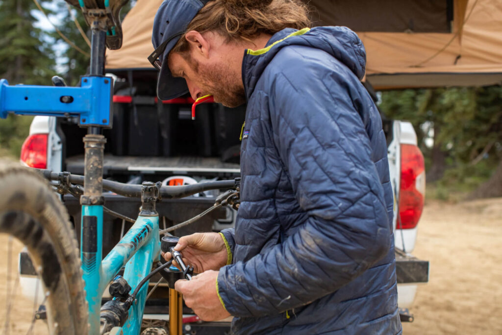 This image shows an individual checking the air pressure in their mountain bike shock. The person is wearing a blue Patagonia jacket and you can see an overland truck in the background.