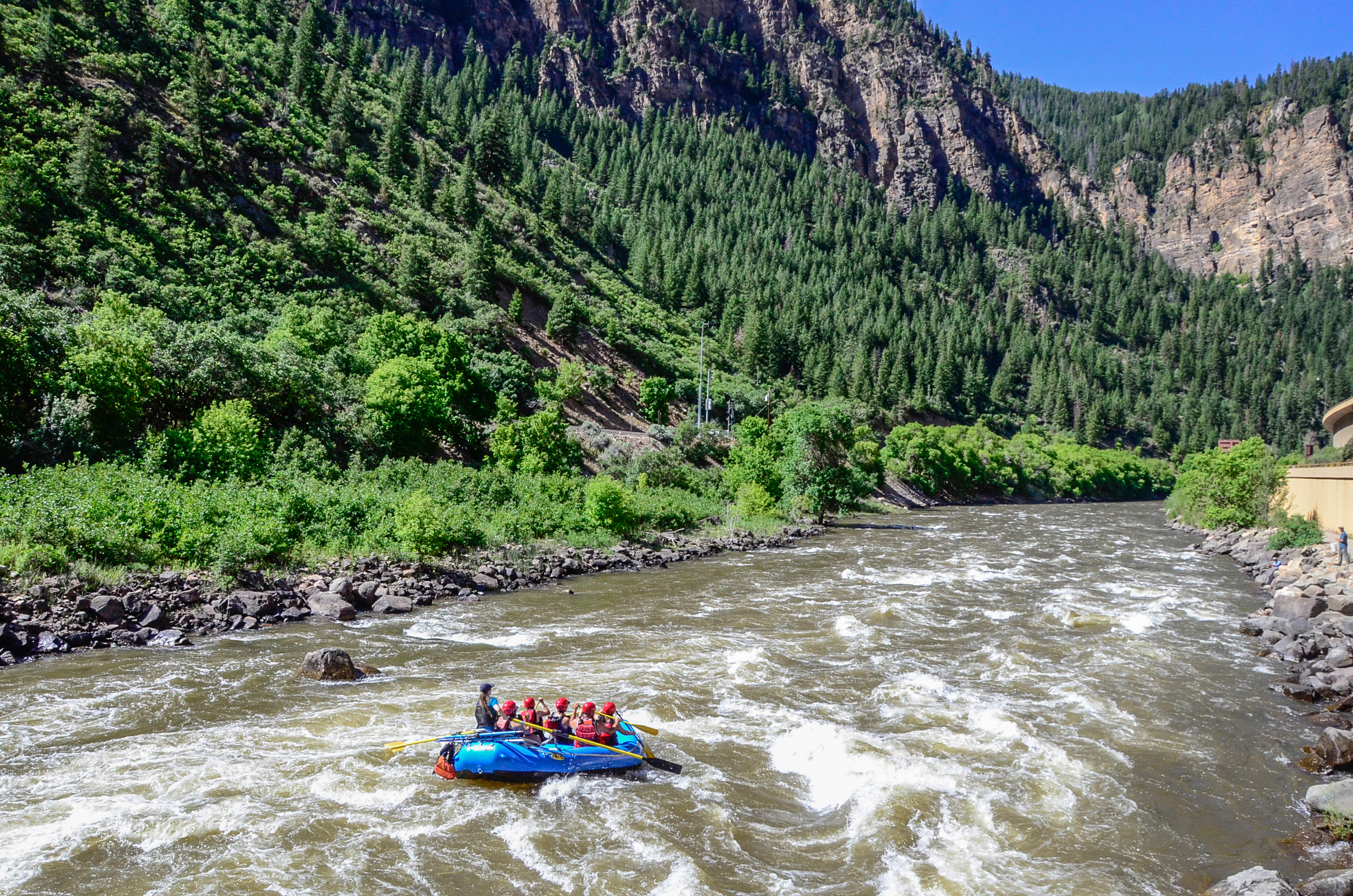 A stunning photo of a full boat gracefully floating down the Colorado River in Glenwood Springs, CO. The canyon is adorned with vibrant green trees and rugged rocks, complemented by the backdrop of a clear, bluebird sky.